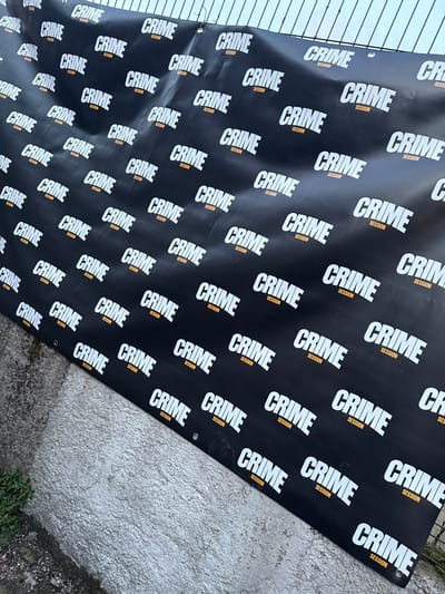 In Noisy-le-Sec, France, a large, black vinyl banner is vertically displayed outdoors. The banner features a repeating pattern of "CRIME" in prominent white, bold, italicized lettering, with "SESSION" in smaller orange text directly below each instance of "CRIME". Metal grommets along the upper edge of the banner secure it to a light-colored, grid-patterned metallic fence, which is visible against a bright, uniform sky. The banner exhibits minor creases. Below the banner, a textured, rough grey concrete surface is visible, along with small patches of green vegetation growing from the ground in the lower left quadrant. No individuals or active subjects are present.
