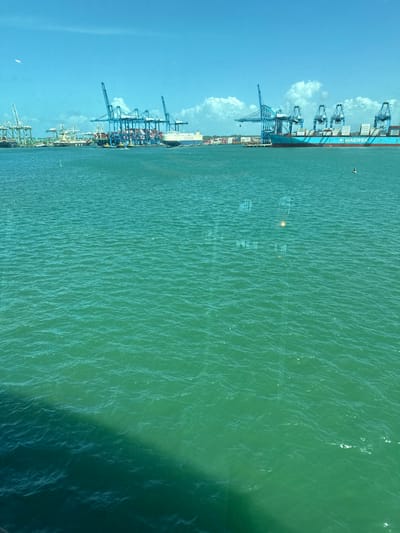 A port scene from above features container cranes and a large container ship along the water's edge. Containers are visible onshore. The water surface shows subtle ripples and reflections, darkening towards the right. A clear blue sky with scattered clouds complements the scene. The image appears to have been taken through a slightly reflective surface.