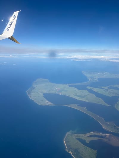 An aerial view of a coastal landscape, likely a peninsula or islands, with a clear blue sky and scattered clouds. The land is predominantly green, suggesting agricultural or natural terrain, with some visible settlements. The deep blue sea surrounds the landmasses. In the distance, a faint line of wind turbines can be seen in the water. The wing of an airplane, with the "RYANAIR" logo visible, occupies the upper left corner of the frame, indicating the perspective is from a flight. The specific location of Kastrup, Denmark, is not explicitly identifiable from the visual information alone.
