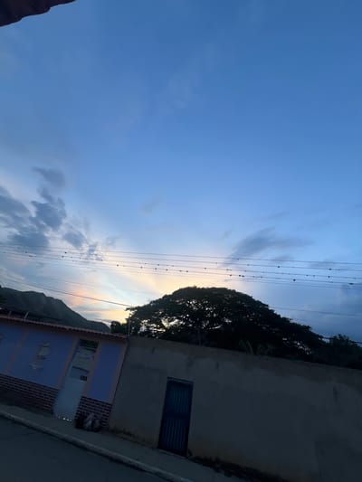 The image shows a street scene in Villa de Cura, Venezuela, at dusk. The sky is a gradient of blue, with patches of clouds and streaks of yellow and pink from the setting sun visible behind a large, silhouetted tree. Several power lines stretch horizontally across the upper portion of the frame, with small, dark objects, possibly birds or debris, perched on them.

In the foreground, a street runs diagonally from the bottom left to the bottom right of the image. On the left side of the street, there is a building with a blue facade and a red brick base. It has a light-colored door and barred windows. Next to it, to the right, is a higher, darker wall with a blue metal gate. The scene appears to be residential, with a somewhat unkempt appearance, as evidenced by some trash bags visible near the blue building. The overall atmosphere is quiet and subdued, characteristic of a village setting at the end of the day. The mountainous landscape is visible in the far background, partially obscured by the tree and the general dimness of the approaching night.