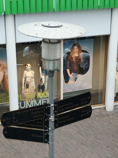 The image shows a street scene in Zaandam, Netherlands, with a focus on a lamppost and signage. The lamppost has a large, circular, weathered metal shade and a ribbed cylindrical body. Attached to the lamppost are several black directional signs.
The signs point to various locations:
- One sign has an information icon and indicates "Toeristeninformatie" (Tourist Information) and "Stadhuis" (City Hall) at 0.1 km. It also indicates "Station" (Station) and "Taxi".
- Another sign points left with "0.1" and an icon that appears to be a building or monument.
- A sign pointing right indicates "0.7" with an icon that looks like a theatre and "0.8" with an icon that resembles a historical building. It lists "Zaantheater" (Zaandam Theater) and "Czaar Peterhuisje" (Tsar Peter House).
- Below these, a sign points left with an arrow and "0.1".
- Another sign points right with an icon that could be a clock or a circle, showing "0.1" and an icon that resembles a building, showing "0.5". It lists "Centrum" (Center) and "Monet Atelier".
- The lowest sign, pointing left with an arrow, reads "OV-fiets" (Public transport bicycle).
In the background, a shop window is visible. The window displays mannequins dressed in clothing. One mannequin wears a plaid skirt and a strapless top, with sunglasses perched on its head and a small bag. Another mannequin is partially visible behind it, wearing denim shorts and a patterned top. A poster with a woman with curly blonde hair and wearing denim shorts is also displayed in the window. The shop appears to be a clothing store, possibly with a "Playing Summer" theme as indicated by large text visible on the window.
The environment appears to be an urban setting with brick paving visible in the foreground. A green, slatted structure is visible at the top of the image, possibly an awning or part of a building.