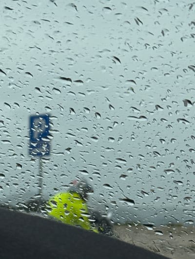 A blurred, wet window obscures the view of a street scene. Raindrops cover the glass. A blue sign is visible on a pole through the raindrops, as is a person wearing a neon yellow garment. A gray structure obscures part of the view. The scene is inferred to be in Aranđelovo, Bosnia and Herzegovina.