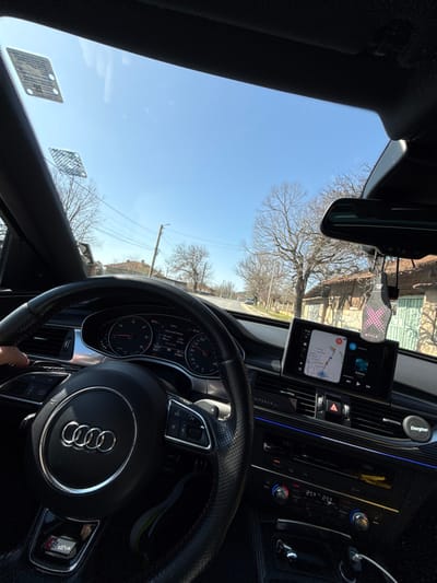 The image depicts the interior of an Audi vehicle, specifically an S-Line model, viewed from the driver's perspective, facing forward on a sunny day in Osenets, Bulgaria.
**Interior Details:**
The driver's left hand is visible on the black leather-wrapped steering wheel, which prominently features the Audi four-ring logo and an 'S line' badge at the bottom. The instrument cluster behind the steering wheel shows two main dials (speedometer on the left, tachometer on the right) with digital readouts between them, including an estimated range of 406 km. To the right of the steering wheel, an infotainment screen displays a navigation map (likely showing a route) on the left side and a music player interface on the right, playing "Rabota". Below the infotainment screen, the car's climate control panel is visible, with digital displays showing "21°C" for both driver and passenger sides. The hazard light button is illuminated. An air freshener, labeled "STYLE" and grey with a pink "X" pattern, hangs from the rearview mirror. Two rectangular stickers are affixed to the upper left portion of the windshield. An Energizer car charger is visible plugged into a port on the passenger side of the center console.
**Exterior Environment (Osenets, Bulgaria):**
The vehicle is situated on a paved road, likely a street within the village. The sky is clear and bright blue, indicating sunny weather. Bare deciduous trees line both sides of the road, suggesting late winter or early spring. On the right side of the road, several older, single-story houses with traditional architecture, featuring brick walls and tiled roofs, are visible. On the left side, a utility pole with multiple power lines runs parallel to the road. Another vehicle is discernible in the distance on the road ahead. The overall scene suggests a quiet, semi-rural setting within Osenets, Bulgaria.