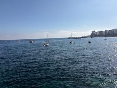 A wide-angle landscape shot captures the serene expanse of Saint Paul's Bay in Malta on a clear, sunny day. The upper half of the image features a bright, cloudless blue sky with only a few faint, wispy clouds near the horizon. The lower half is dominated by the calm, dark blue waters of the bay, which show gentle ripples and reflect sparkling sunlight, particularly on the right side of the frame.
Numerous boats are dispersed across the water. On the left, a white motorboat with a red trim moves away, leaving a white wake behind it. Towards the center, a sailboat with a tall mast is anchored, and several smaller boats and dinghies are also visible, some appearing to be moored. Further towards the right, closer to the distant shore, there's a larger white boat or small ship, possibly near a pier structure that extends from the land. Lines of red and white buoys are systematically arranged across the water, likely delineating swimming zones or mooring spots.
Along the distant right shoreline, a cluster of modern, multi-story buildings, predominantly white and grey, forms a coastal cityscape, suggesting a developed area. The bright conditions and clear visibility indicate either morning or early afternoon. The overall scene evokes a peaceful and leisurely atmosphere typical of a Mediterranean coastal area. No discernible text is visible within the image.