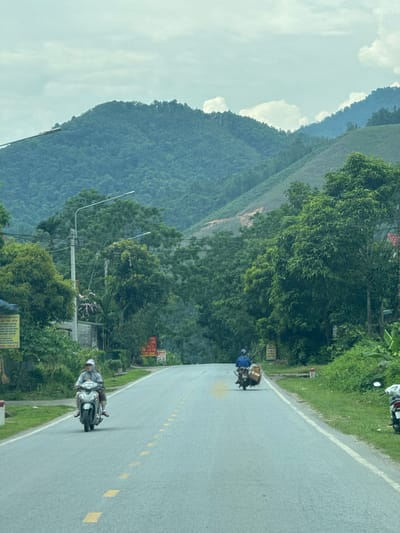 The image shows a paved road cutting through a landscape of lush green mountains under a cloudy sky. On the road, two individuals are riding motorbikes in the same direction. The motorbike in the foreground carries two people and the one behind appears to be carrying some cargo. The road has yellow dotted lines marking the center. On the side of the road are trees, vegetation, and utility poles. Based on the road signage, foliage, and mountainous terrain, the location appears to be along a road in the city of Hà Giang 2 Ward, Vietnam.