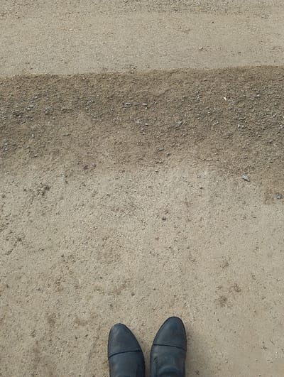 The image depicts a top-down view of an individual's feet standing on an outdoor surface in Qanlikul, Uzbekistan. A pair of dark-colored, possibly black or dark grey, low-cut shoes are positioned at the bottom center of the frame. The ground immediately in front of the shoes is composed of light brown, compacted soil or fine dirt with minimal debris. Approximately one-third of the way up from the bottom, the ground transitions horizontally to a darker, more textured surface characterized by a higher concentration of small stones and gravel. This coarser section extends for a significant portion before transitioning back to a lighter, less gravelly texture in the upper background. No other individuals, objects, or actions are evident.