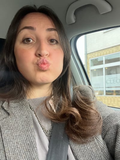 A female individual is depicted in a selfie, seated inside a vehicle. She has long, dark brown hair, brown eyes, and fair skin, with visible blush on her cheeks. Her lips are pursed into a pouting expression, and she is looking directly at the camera. She wears a grey t-shirt under a grey tweed-patterned jacket. A silver-colored chain necklace is visible around her neck, and a black seatbelt crosses diagonally over her chest. The vehicle's light grey interior grab handle is visible in the upper right. Through the passenger-side window, a light-beige textured building is visible. A sign on one of the building's windows partially displays the text "Sonnenstudio CLEO". This building is located at Obergruppenbacher Str. 12, in Untergruppenbach, Germany.