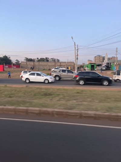 The image presents an outdoor urban scene in Biashara ward, Kenya, observed from a low angle across a grassy median strip. A multi-lane paved road occupies the mid-ground, featuring vehicular traffic. From left to right, a white sedan, a light-colored pickup truck, and a black SUV are in motion, followed by a white tuk-tuk partially visible on the far right. On the left dirt verge beside the road, a male individual in a blue shirt and dark pants walks rightward. In the background, a light-brown brick wall spans horizontally, behind which various individuals are visible. Further back, a red billboard displaying the 'Airtel' logo is present on the left. Multiple residential and commercial buildings are visible, including structures with rooftop water tanks, a cluster of green-painted stalls, and a building under construction adjacent to a structure with a dome and minaret. Utility poles equipped with overhead power lines and a street light fixture punctuate the background against a clear, light blue sky.
