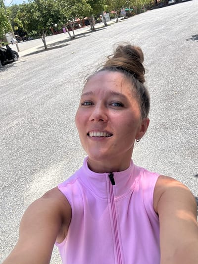 A young woman with her hair in a messy bun smiles for a selfie outdoors. She is wearing a bright pink sleeveless athletic top with a zippered collar. Her skin is flushed, and she has a small nose ring and hoop earrings. The background is a dusty, gravel-covered parking lot with several trees and buildings visible in the distance. The scene appears to be in Thung Yai, Thailand, suggesting a warm, outdoor environment.