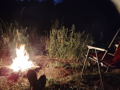 This is a nighttime, first-person perspective photograph of a campfire scene. In the foreground, two feet, likely belonging to the photographer, are visible with dark shoes or socks, placed on the ground. To the left, a bright, active campfire is burning, illuminating the surrounding dry grass and dirt. In the middle ground, tall, green reeds or grasses grow densely. To the right, a red camping chair with a metal frame is set up, partially visible. The background is dark, with the silhouettes of trees and a sliver of a dark blue night sky. A faint, curved white line, possibly a streak of light or a star trail, is visible in the upper portion of the sky. The setting appears to be a natural, outdoor area, possibly a campsite near a body of water, given the vegetation and the open space. The location is Balyko-Shchuchynka, Ukraine.