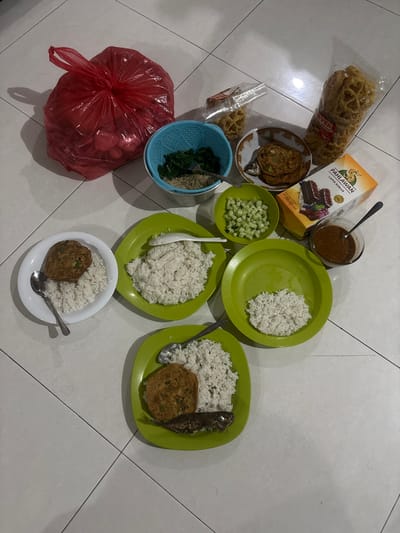 A top-down, eye-level shot shows a meal laid out on a tiled floor, likely in Kalijaten, Indonesia. The meal consists of three plates of white rice, each accompanied by a fried patty and a small fried fish on two of the plates. There are also side dishes in small bowls: chopped cucumber, cooked leafy green vegetables with bean sprouts, and a small dish of dark sauce. Two bags of crunchy snacks are visible, along with a box of "Pahlawan Lapis Kukus" cake. A large red plastic bag, possibly containing more food or groceries, sits to the left. The overall scene suggests a family meal being prepared or consumed on the floor.