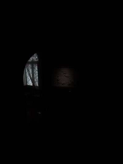 The image captures a dimly lit interior, possibly in Sintra, Portugal. A partially visible arched window with lace curtains is positioned to the left, allowing some light to enter the room. Adjacent to the window, a chair is silhouetted against the dark backdrop. Further to the right, a wall features a relief or mural, faintly illuminated. The overall environment is dark.