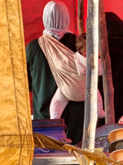 A figure wrapped in fabric is partially visible. The figure has a grey head covering, a light-colored sling, and is clothed in dark green. Rough wooden poles cross the image, obscuring parts of the figure. Miscellaneous objects including what seem like spoons, and a blue and red object are visible in the background. The foreground is dominated by a yellow textured surface. Red fabric is seen behind the figure.