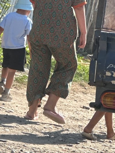 The image shows a street scene in the city of International, Kyrgyzstan. In the foreground, the lower bodies of three people are visible. A child in a light blue bucket hat and white t-shirt walks to the left, wearing black shorts and grey socks. To their right, a person in a patterned jumpsuit with green and red paisley designs walks forward, wearing pink slip-on shoes. A second person, further to the right, walks with their back to the camera, wearing light-colored sandals. They are partially obscured by a dark blue vehicle with a portion of a red taillight visible on the right side of the frame. The ground is a mixture of dirt, gravel, and small stones, with patches of dry grass and vegetation visible in the background. The lighting suggests it is daytime, with strong shadows cast on the ground.
