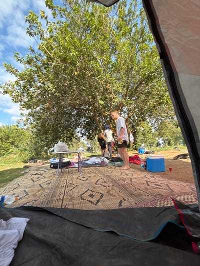 The scene depicts an outdoor camping or picnic area, viewed from within a dark-colored tent. A large, patterned woven mat, predominantly beige with dark brown and red geometric designs, covers the ground immediately outside the tent opening.
Three individuals are present. A fair-skinned male, barefoot, wearing a white short-sleeved shirt and dark shorts, stands upright on the mat towards the right side of the frame, holding a yellow beverage can in his right hand. He appears to be looking slightly left. Behind him and to his left, two other individuals are partially visible. One person, wearing dark clothing, is bent over, seemingly attending to items on the ground. The third person, wearing lighter clothing and a blue head covering, is partially obscured behind the first and appears to be standing.
Various camping items are scattered on and around the mat. To the left, a small, collapsible table with purple legs holds a white plastic bag. Nearby, a grey sleeping pad or mat is laid out, surrounded by folded clothing and bags. To the right, a blue cooler is visible on the bare earth adjacent to the mat, with two red plastic cups and other miscellaneous gear.
A large tree with dense green foliage provides shade over the area, its branches extending across the upper portion of the image. The ground beneath the tree is primarily exposed brown earth. To the far left, beyond the mat, a patch of green grass transitions into dense green vegetation. The sky above is clear with scattered white clouds, indicating daylight. The location is in the Galil Elyon Regional Council, Israel.