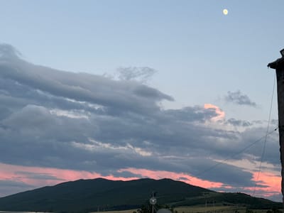 The image captures a dusky sky over a hilly landscape in Mrgvali Chala, Georgia. A crescent moon is visible in the upper right quadrant of the sky. The sky transitions from a pale blue at the top to soft pink and gray hues near the horizon, indicative of either sunrise or sunset. Dark, layered clouds stretch across the upper half of the frame, with some lighter, wispy clouds interspersed.

Below the sky, a dark, verdant mountain dominates the lower portion of the image, its slopes covered in what appears to be dense vegetation. The terrain suggests a rural or natural setting. In the foreground, along the right edge, a weathered stone structure or wall with a dark, angular object perched on top is visible, possibly a rooftop or antenna. A thin wire runs diagonally across the frame, suggesting human infrastructure amidst the natural scenery. The overall mood is serene and tranquil, with the soft lighting and the presence of the moon contributing to a peaceful atmosphere. The time of day is clearly twilight.