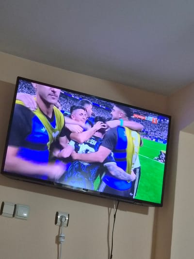 The image displays a television screen showing a group of male soccer players celebrating on a green field, likely during a match. The players are wearing blue and yellow jerseys. One player in the foreground has his arms around another player who is smiling. A scoreboard is visible in the background with text that appears to be in Cyrillic and numbers indicating a score of 1-0. The television is mounted on a light-colored wall, below which are two white light switches and a white electrical outlet with a plug and cord. The scene is set in an indoor environment, possibly a living room, in the city of Kaleytsa, Bulgaria.