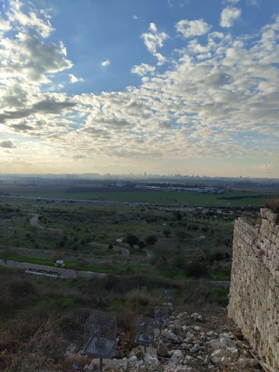 The image, taken in landscape orientation, presents a panoramic view from an elevated position, likely atop ancient ruins or a hill, overlooking a broad valley and distant cityscape.

In the immediate foreground, on the right side, a substantial stone wall constructed from rough-hewn, light-colored stones dominates. Below the wall, the ground is covered with rubble and loose stones, interspersed with dry vegetation. Several square, metal cage-like structures are visible among the debris.

The midground transitions into a wide, verdant expanse of fields and uneven terrain, characterized by patches of green grass and scattered low shrubs and trees. Faint pathways or dirt roads can be discerned winding through this landscape.

On the far left, a hazy silhouette of a distant city or town stretches across the horizon, with numerous buildings just visible through the atmospheric haze.

The upper portion of the image is filled with a vast, partly cloudy blue sky. Numerous white, fluffy clouds are scattered across the frame, some appearing brighter than others, suggesting sunlight breaking through. The overall lighting implies either early morning or late afternoon, with a soft, diffused glow.

There is no specific activity or event taking place; the scene is a static landscape. The weather appears clear and bright, with pleasant cloud cover. The presence of ancient-looking stone structures overlooking agricultural land and a modern city is consistent with the context of Rosh HaAyin, Israel, a region known for its historical sites like Tel Afek/Antipatris, offering views of the surrounding plains and contemporary development. No visible text is present in the image.