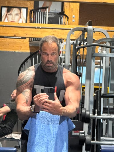 A medium shot shows a muscular man in a gym setting, likely located in Jemelle, Belgium. He is holding a smartphone and appears to be taking a selfie or looking at something on the screen. He has graying hair, a beard, and tattoos on his right arm. He wears a black tank top and a gold bracelet on his right wrist, with a watch on his left. A blue towel is draped around his lower body. Gym equipment is visible in the background, including a weight machine. A wooden wall with posters or framed images is also present. The overall lighting appears bright and possibly artificial.