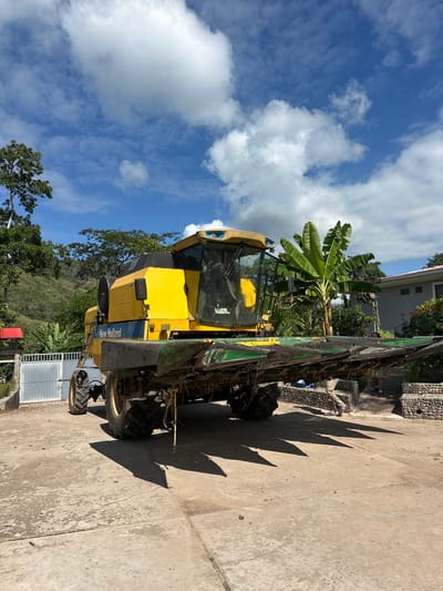 The image depicts a yellow New Holland combine harvester parked on a concrete surface, likely a driveway or yard. The machine has a large harvesting head at the front. In the background, lush green vegetation, including banana trees and a hillside covered in trees, suggests a rural or semi-rural environment. A partially visible building with a grey facade and a red-roofed structure are also present. The sky is blue with scattered white clouds. There are two individuals visible in the background, one partially obscured by the combine. The scene does not appear to be in the city of Buenos Aires, Peru, as there are no recognizable landmarks or urban features consistent with that location.