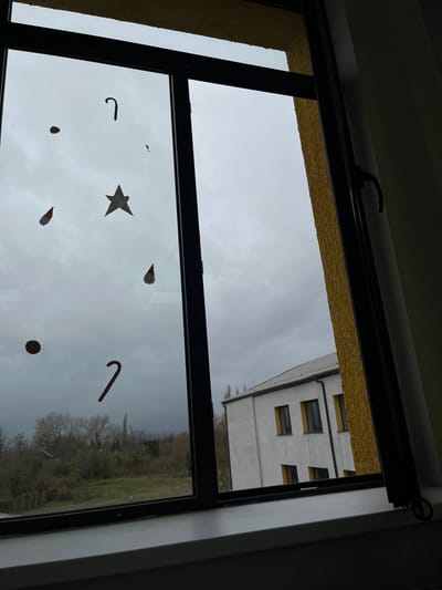 The image presents an exterior scene viewed through a window from an interior vantage point in ლესის თემი, Georgia. The window features dark frames, with the right pane partially open inwards, revealing a section of an adjacent exterior wall finished with a yellow, textured stucco. Adhered to the window glass are six decorative decals: two dark, curved candy cane shapes; one yellow five-pointed star; two circular figures depicted wearing red pointed hats; one red and green striped spherical object; and one small, dark, circular object. Beyond the window, a two-story white building with a light-colored tiled roof and multiple rectangular windows with dark frames and yellow sills is visible on the mid-ground right. To the left and in the distance, an expanse of bare deciduous trees and green ground cover is discernible under a uniformly overcast and grey sky. No individuals or active interactions are present within the frame.