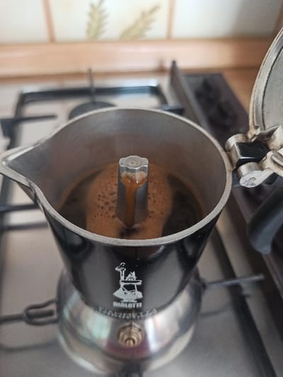 An aluminum and black-enameled Bialetti Moka pot is positioned on a gas stovetop within a kitchen in San Marzano sul Sarno, Italy. The device is actively brewing coffee, evidenced by the dark liquid and light brown crema emerging from the central column into the open upper chamber. The Moka pot's lid is hinged open towards the right. The stovetop features metal grates. In the blurred background, a tiled backsplash with a stylized light-colored foliage pattern is visible above a light-colored counter or cabinet. No individuals are present in the frame.
