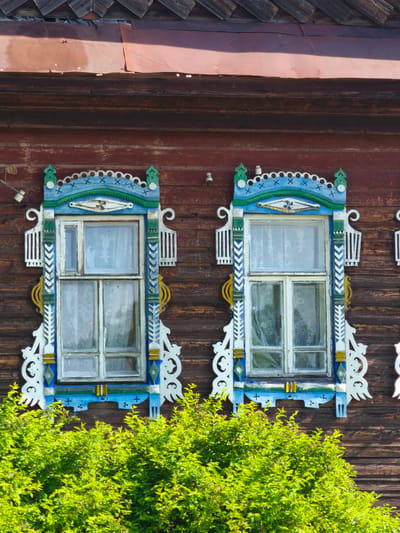 A close-up, medium shot captures the detail of two ornately carved wooden window frames on the exterior of a weathered, dark brown wooden building in the city of Zhukovo, Russia. The windows themselves are made of multiple panes with white curtains visible inside, and the wooden frames are painted in a vibrant blue, green, and white color scheme, featuring intricate scrollwork, decorative patterns, and small carved finials at the top. Below the windows, a dense, bright green bush fills the lower portion of the frame, its leaves catching the sunlight. The building's roof, made of dark, overlapping wooden shingles, is visible at the top, with a section of reddish, corrugated metal sheeting covering part of the upper wall. The overall scene is static and focused on the architectural details and the surrounding greenery.