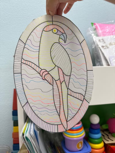A person's hand holds up an oval-shaped piece of paper with a black-outlined drawing of a parrot on it. The parrot is colored with yellow, green, orange, red, and blue lines. The background is a light blue wall. In the lower right corner, there are out-of-focus images of a stack of colorful toys and some papers. The scene is indoors and appears to be in a domestic setting.