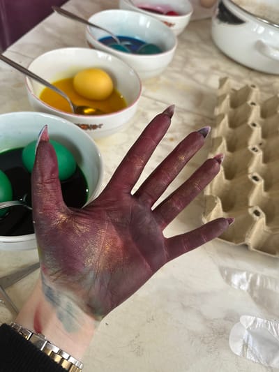 A person's hand, stained purple, gold, green, and blue, is positioned above bowls of egg dye. The nails have clear tips. Bowls of dye in red, blue, yellow, and green are visible. A silver spoon is in each bowl. An egg carton and a disposable glove are in the background. The wrist has a silver wristwatch with a white face. The location is most likely an indoor space in a residential area. There is no visual information in the image to suggest that this event took place in Galiche, Bulgaria.