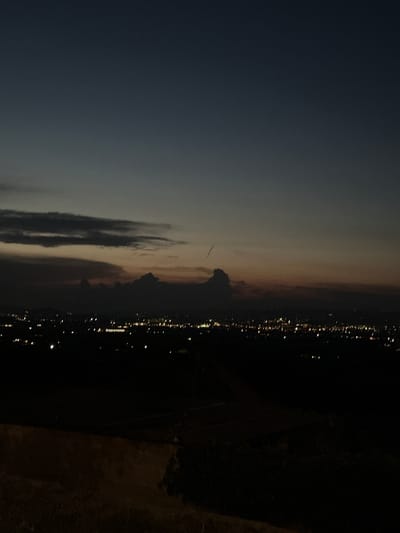 This image captures a twilight view over the city of Cannara, Italy, from an elevated vantage point. The sky transitions from a deep, dark blue at the top to lighter shades of grey and dusky orange near the horizon, indicating either dawn or dusk. A few dark, wispy clouds are visible in the sky. Below the sky, the silhouette of Cannara is spread out, with numerous small, yellow lights indicating the presence of buildings and streetlights. The foreground consists of dark, indistinct terrain, possibly hills or fields, with the texture suggesting natural landscape. There are no visible people, vehicles, or specific landmarks that identify the exact location within Cannara, but the overall scene depicts a city at night or during twilight.