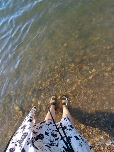 The image shows a person's legs and feet submerged in shallow, clear water at the edge of a body of water. The ground beneath the water is composed of pebbles and rocks of various sizes and colors. The person is wearing black sandals with silver buckles and a white dress with a black abstract floral pattern. A faint tattoo is visible on the person's ankle. The water has gentle ripples on its surface. The scene appears to be a natural shoreline, potentially in Oryahovo, Bulgaria, given the context. The lighting suggests it is daytime.