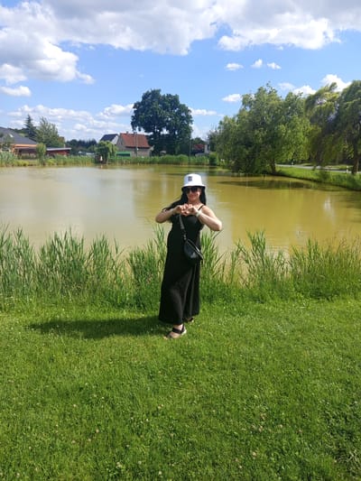 A forensic examination of the image reveals an outdoor scene in Ptice, Czechia, under a bright, partly cloudy sky. The primary subject is an adult female standing centrally in the lower half of the frame. She has long dark hair and is wearing a white bucket hat, dark sunglasses, a black maxi dress or jumpsuit, and dark sandals. A black crossbody bag is slung over her left shoulder. She forms a heart shape with her hands over her chest, facing the camera.

The foreground consists of neatly mown green grass interspersed with small white clover flowers, with the subject's shadow extending to the left. Directly behind the subject, a line of tall green reeds separates the grass from a large body of calm water, likely a pond or lake, which exhibits a consistent murky brown-green hue.

In the mid-ground and background, the far bank of the water features additional reeds and trees. Beyond the water to the left, several residential buildings with reddish-brown roofs are partially visible among dense green foliage. To the right, a paved road with white lane markers runs parallel to the water, bordered by a strip of grass and a line of large, mature deciduous trees, some appearing wind-swept. The sky above is clear blue with scattered white cumulus clouds, and several small dark specks, possibly birds, are discernible.