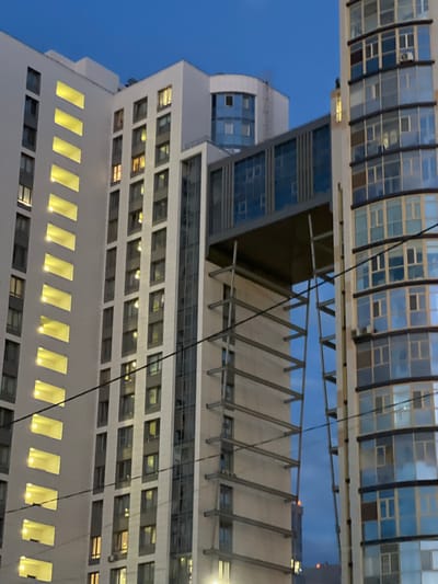The image shows a contemporary multi-story residential building at dusk, with a clear blue sky above. The building features a light-colored facade with numerous windows, some of which are illuminated with a warm yellow light, suggesting interior lighting. A prominent architectural feature is a glass-enclosed walkway connecting two sections of the building, supported by a metal structure. To the right, another building with a facade of blue-tinted windows is visible. The setting appears to be urban, with electrical wires crossing the frame, hinting at power distribution infrastructure. The location is identified as Dachnoe, Russia. There are no people or animals visible in the frame. The overall scene conveys a sense of modern residential architecture in a twilight urban environment.