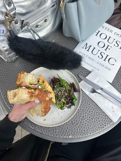 The scene depicts an outdoor cafe table at Serpentine South Gallery in Kensington Gardens, City of Westminster, United Kingdom. A person's left hand, adorned with light-colored nail polish, holds a half-eaten sandwich, consistent with a baguette or ciabatta, containing ham, pesto, and a reddish spread. A white ceramic plate next to the sandwich holds a side salad composed of mixed greens, sunflower seeds, and chia seeds. A fork and knife rest on a folded white paper napkin. To the left, a silver metallic handbag with a "Sick" charm is visible, partially obscured by a black faux fur object. Behind these, a light blue leather handbag is positioned. A white pamphlet titled "PETER DO HOUSE OF MUSIC" for an exhibition running "10 OCT 2025 – 8 FEB 2026" at "SERPENTINE SOUTH" lies on the table. The table surface is dark grey/silver with a dimpled texture. The person's lower torso, clad in dark clothing, is partially visible.