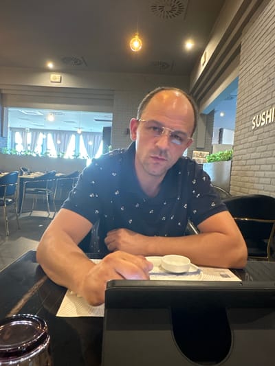 A man with a shaved head and glasses sits at a table in a restaurant. He is wearing a dark blue polo shirt with a white floral pattern. His arms are resting on the table, and his hands are in front of him. There is a small white bowl on the table in front of his hands. To his left, a dark-colored glass is visible. In the background, there are other tables and chairs, as well as a brick wall with the word "SUSHI" written on it. The lighting suggests it is indoors, possibly in the city of Ponteterra, Italy, given the context of the prompt.