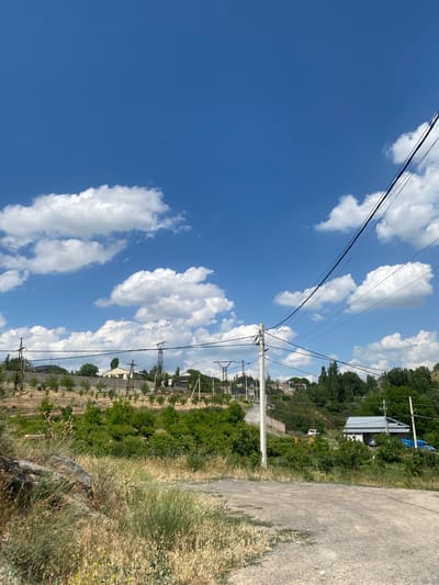This media file captures a bright, sunny day in Ptghni, Armenia. The scene is dominated by a vibrant blue sky filled with fluffy white clouds, suggesting fair weather. In the foreground, a rough, unpaved road stretches diagonally across the right side of the frame, bordered by dry, tall grass and some vegetation.
A prominent feature of the image is a series of power lines and utility poles, crisscrossing the sky and extending into the distance. These lines suggest the presence of human infrastructure in a semi-rural or suburban setting. Beyond the road and vegetation, a gently sloping hillside is visible, dotted with trees and scattered buildings. Some of these structures appear to be houses, while others might be sheds or workshops, featuring metal roofs and simple designs. Further in the background, a dense line of trees marks the top of the hill, and in the far distance, more buildings are faintly visible, possibly indicating a small village or community.
The overall impression is one of a warm, pleasant day in a rural or semi-rural Armenian landscape. The lighting suggests it is likely late morning or early afternoon, given the bright sunlight and clear skies. There is no discernible text in the image. The scene is peaceful and conveys a sense of everyday life in this location.
