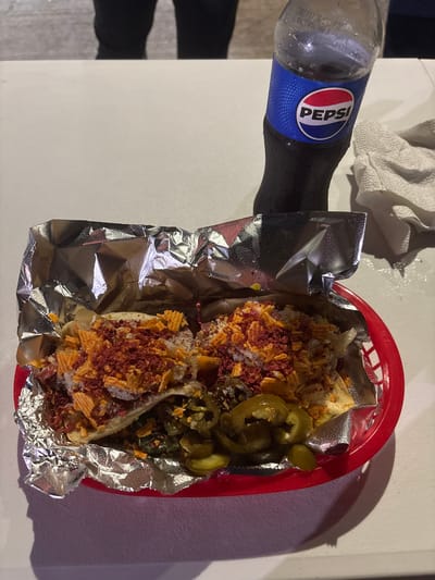 A top-down shot shows a meal in Tampico, Mexico. A small, glass Pepsi bottle lies horizontally to the left of a red plastic basket lined with foil. Inside the basket are two corn tortillas topped with yellow chips, crumbled red meat, a white powder, and jalapeños. A white napkin sits above the bottle. The scene appears to be on a white table. A shadowy figure is partially visible on the left edge.