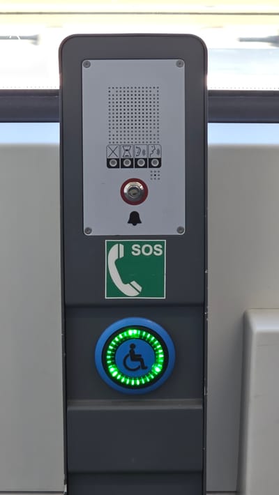 This is a close-up, eye-level, outdoor shot of a public communication panel, likely on a tram or bus in Neu-Ulm, Germany. The panel is dark grey and mounted vertically. At the top is a silver plate with a speaker grille. Below the grille are four circular buttons with icons: a crossed-out 'X', an hourglass, a person with a hearing aid, and a person speaking. Below these buttons is a red emergency button with a silver center. Below that is a bell icon and the text "SOS" on a green background with a white telephone symbol. The bottom of the panel features a blue circular button with a green illuminated ring of lights and a black wheelchair symbol in the center. The background shows a blurred exterior with bright daylight, suggesting the panel is located near a window or open area.