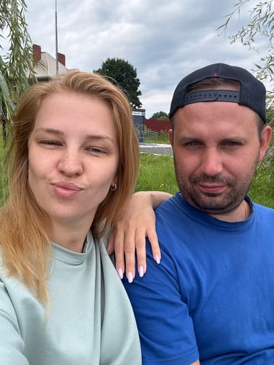 In Breytovo, Russia, a close-up, waist-up selfie captures a couple outdoors under a cloudy sky. The woman, positioned on the left, has light blonde hair and is wearing a light teal sweatshirt. She is making a puckered-lip expression, looking towards the camera, with her left hand resting on the man's shoulder. Her right arm is not visible. She has light skin with freckles and is wearing small hoop earrings.

The man, on the right, is wearing a blue t-shirt and a dark baseball cap. He has short, dark hair and stubble. He is looking directly at the camera with a neutral expression. A gold chain necklace is visible around his neck. His left arm is around the woman's shoulder, with his hand resting on her back.

Behind the couple, a building with a red-tiled roof and a brick chimney is partially visible to the left, with some greenery and trees in the background. A sign with Russian text is visible on a pole to the right of the building. The ground is covered in grass.