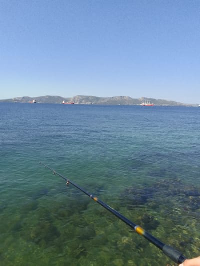 The image depicts a serene scene by the sea, likely in the Municipal Unit of Elefsina, Greece. In the foreground, a person's hand holds a fishing rod, casting a line into the clear, shallow waters. The water itself is a striking turquoise, revealing the rocky seabed beneath. The fishing rod, black with golden accents and a black grip, extends into the frame, its line disappearing into the water.

In the middle ground, the deep blue sea stretches towards a distant, hazy shoreline. Several large cargo ships are visible on the water, suggesting a maritime or industrial area. One ship, painted red and white, stands out with its distinctive silhouette. The background is dominated by a range of low, arid hills that form the coastline. The sky above is a vast expanse of clear, bright blue, indicative of a sunny day.

The time of day appears to be late morning or early afternoon, given the high sun and clear visibility. The weather is fair and calm, with only gentle ripples on the water's surface. The overall mood is one of tranquility and peaceful recreation. There is no visible text in the image.