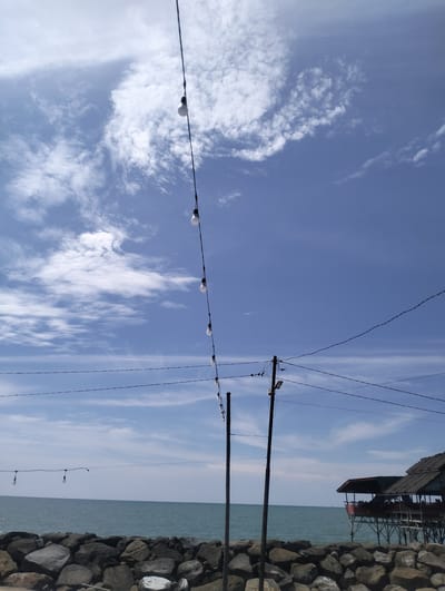 The image depicts an outdoor scene at the beach in Lancok, Indonesia, under a blue sky with scattered white clouds. In the foreground, a wall of large, dark rocks lines the edge of the water. Beyond the rocks, the sea stretches out to the horizon. A few string lights with bare bulbs are suspended on wires above the water, with one set extending from the left and another from the right side of the frame. Two tall, dark poles with wires are also visible, one to the right of the center and another further to the left. On the right side of the image, a wooden stilted structure with a thatched roof, resembling a small restaurant or cafe, is situated over the water, with several people visible inside. The overall atmosphere is bright and summery.