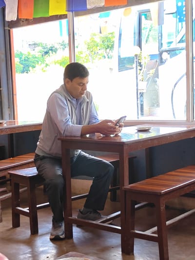 A man sits at a wooden table in a cafe with large windows overlooking a tree-lined street. He is wearing a light blue collared shirt and a grey vest. He is holding a smartphone in his hands and appears to be looking at it. A small, empty plate sits on the table in front of him. The cafe has wooden benches and tables. Prayer flags are hanging from the ceiling. The scene is lit by natural light coming from the windows. The location is the city of Tharpu, Nepal.