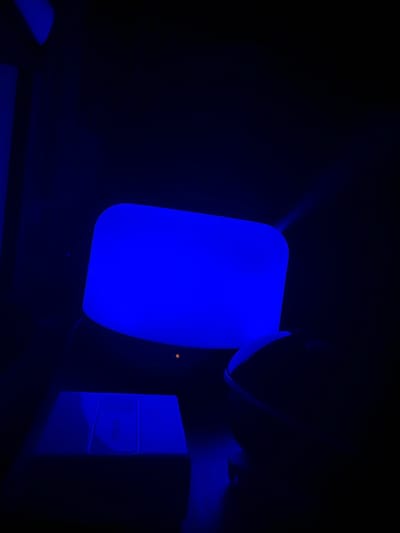 The image captures a close-up, low-light view of a desk with a bright blue illuminated object, possibly a humidifier or a modern lamp. To the right of the blue object is a dark-colored computer mouse. In the foreground, a rectangular object, likely a scale, is also illuminated with a faint blue light and shows some subtle markings or a screen. The background is almost entirely dark, with the blue light casting a glow on the surrounding surfaces. There are no people visible in the image. The scene appears to be indoors, likely in a personal workspace or bedroom. The image was taken in McMasterville, Canada.