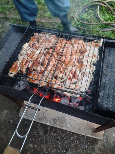 In Il'bukhtino, Russia, a close-up shot captures chicken pieces and sliced onions grilling over glowing red coals in a black metal barbecue grill. The grill is situated outdoors on a weathered concrete surface. The background shows green grass and a coiled garden hose, with the lower legs of a person wearing blue jeans and black boots visible in the upper left portion of the frame.