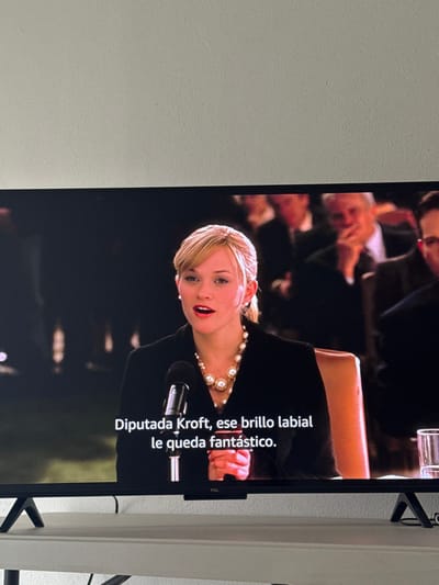 The image shows a blonde woman with a pearl necklace speaking at a microphone. She is wearing a black top. The background is blurred, showing other people seated behind her. Spanish subtitles appear on the screen, reading "Diputada Kroft, ese brillo labial le queda fantástico." This image appears on a TCL television. The wall is painted off-white.