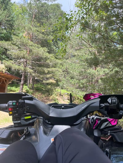 The image shows a first-person perspective of someone seated on a black ATV, parked in a wooded area. The handlebars and control panel of the ATV are in the foreground, with a helmet, likely pink and grey, placed on the ATV in front of the seated person. The background consists of a dense forest with tall pine trees and other green foliage. A small wooden structure, possibly a shelter or shed, is visible to the left. The lighting suggests it is daytime. The location is identified as Simeonovets, Bulgaria.
