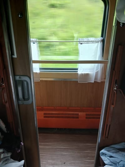 This is a photograph taken from inside a train carriage, looking out through an open doorway towards the window. The scene is one of movement, as indicated by the blurred green foliage seen through the window, suggesting the train is traveling through a natural landscape, likely rural or mountainous. The interior of the carriage is simple and functional, with wooden paneling on the walls and a wooden floor. A gray handle is visible on the left doorframe, and a brown handle on the right. An orange heating unit with a slatted front is mounted near the floor beneath the window. Two sheer white curtains hang on a rod across the lower part of the window. On the left, near the open doorway, a pile of items including dark fabric and a pink cloth are visible. On the right, a folded light blue fabric is present. The overall impression is that of a basic, utilitarian train compartment. The image does not contain any elements that would specifically place it in Krasnaya Polyana, Russia.
