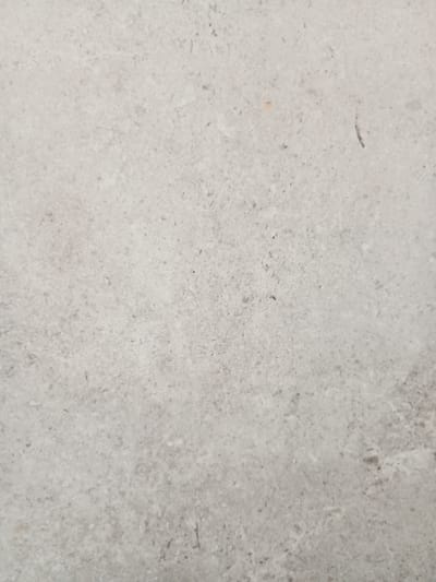 The image displays a close-up, textured view of a light gray, mottled surface, likely concrete or stone. There are no discernible subjects like people, objects, or a clear scene. The activity, time of day, weather, or location cues are not apparent. The texture of the surface is characterized by subtle variations in color and small, dark speckles scattered across the lighter gray background. One hint of color appears as a very small, faint orange mark on the left side of the image. The overall impression is of a neutral, possibly weathered, outdoor or industrial surface. No text is visible.