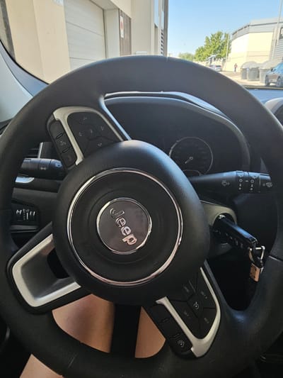 The image shows a close-up view of a black Jeep steering wheel, with the Jeep logo prominently displayed in the center. The steering wheel has various buttons on either side, including controls for the audio system, cruise control, and potentially other vehicle functions. A car key is inserted into the ignition on the right side of the steering column. The background is slightly out of focus, but suggests an outdoor setting with a building and trees visible. Given the context of the location, it is highly probable that this image was taken within the city of Cuarte de Huerva, Spain.