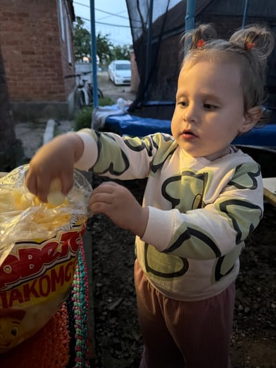 A young child, likely a toddler, is reaching into a clear plastic bag. The bag appears to contain some kind of food product, possibly snacks, with text visible on its red and yellow packaging. The child is wearing a light-colored long-sleeved shirt with a green and black pattern and pink ribbed pants. Their hair is styled in two small buns. The setting appears to be outdoors, possibly a backyard or an area near a building. In the background, a brick building, a bicycle, a white car, and a trampoline structure are visible, suggesting a residential area in Komsomolskiy, Russia. The lighting suggests it may be dusk or early evening.